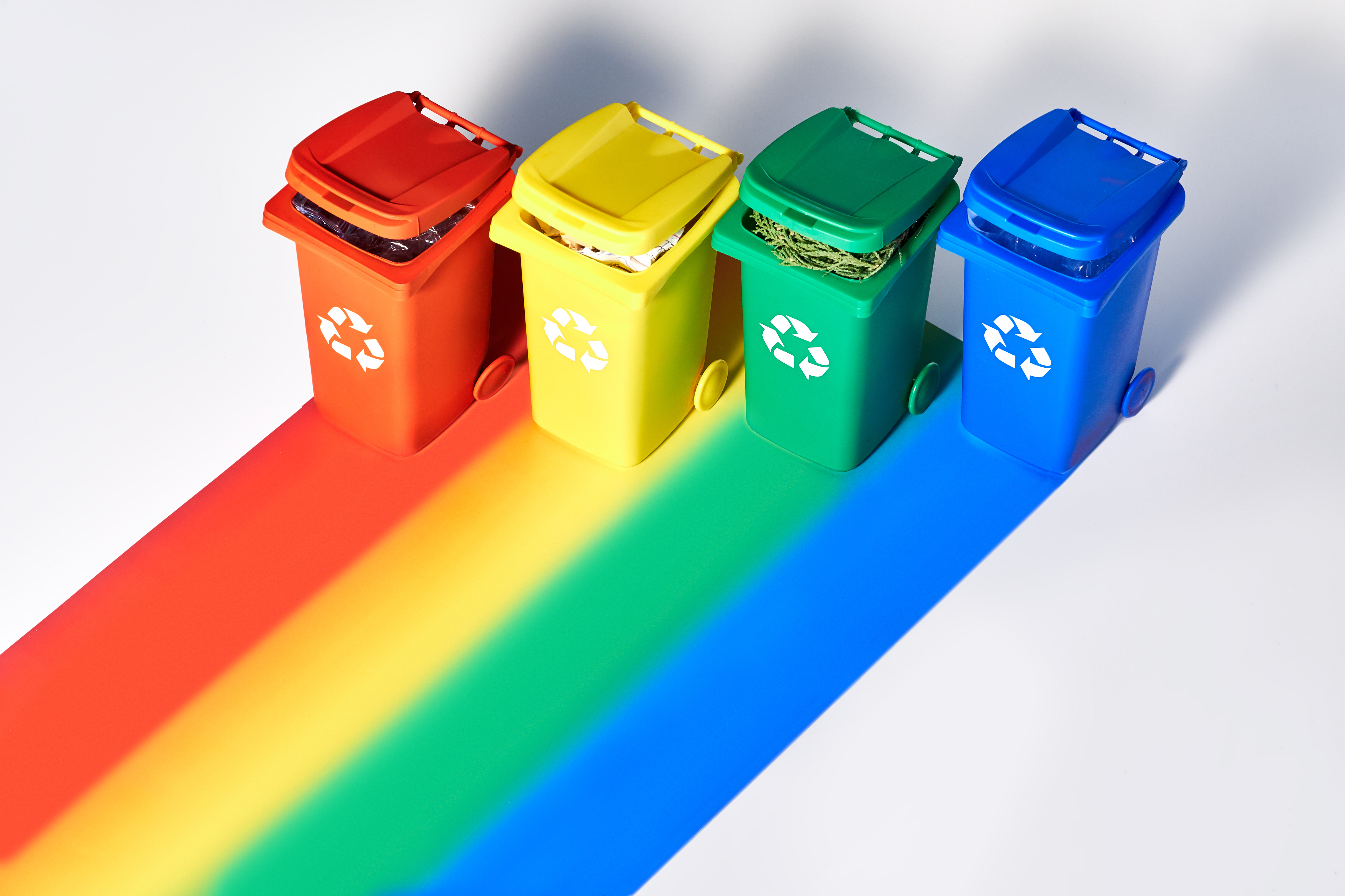 Four color coded recycle bins, isometric projection on geometric rainbow paper background with copy-space. Recycling sign on the bins - red, blue, yellow, green. Waste separation concept background.