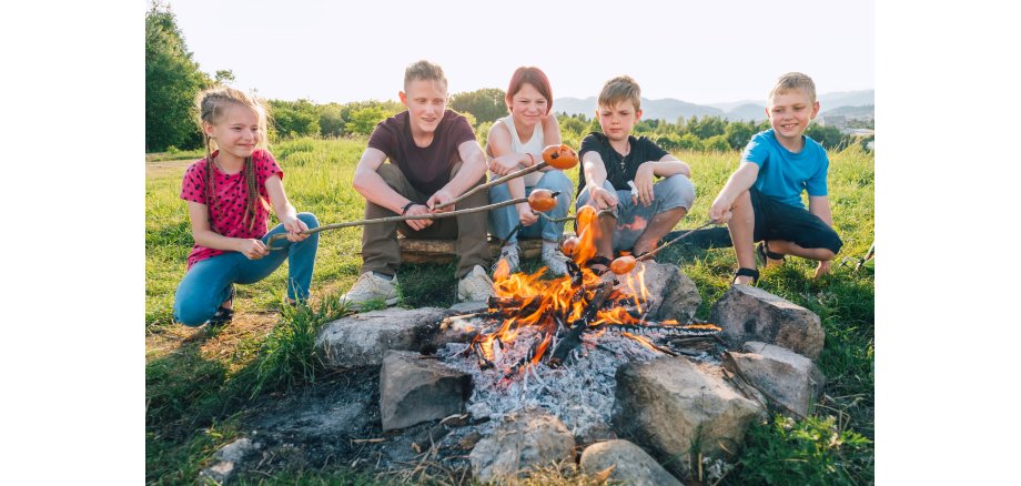 Group of Kids - Boys and girls cheerfully smiling and roasting sausages on long sticks over a campfire flame. Outdoor active time spending or camping in Nature concept.