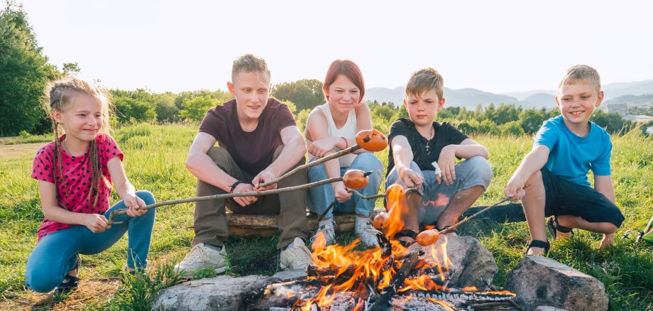 Group of Kids - Boys and girls cheerfully smiling and roasting sausages on long sticks over a campfire flame. Outdoor active time spending or camping in Nature concept.